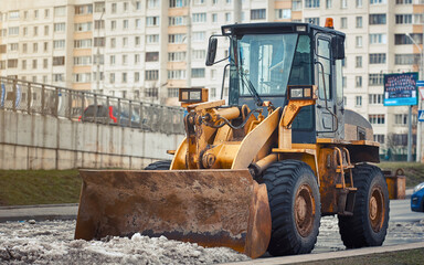 Yellow front loader with snow plow blade removing snow. Snow removal machine for clearing parking lot after winter storm. Loader bucket removing snowdrift. Tractor clean snow, removal road services