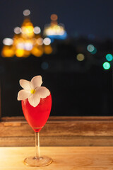 Glasses of red cocktail with ice and white flower on wooden table front of windows in the night with beautiful light blurred bokeh background.