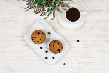 Cupcakes with black currants on a white wooden cutting board against the background of a cup of tea. View from above.