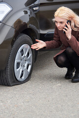 A young blonde woman talking on a mobile phone near her car with a flat tire.