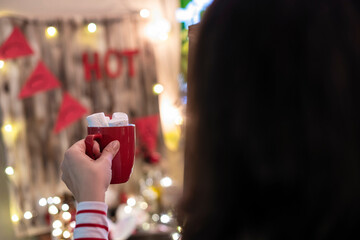 Woman hand with cup of hot cocoa with marshmallows at christmas time opposite small home cocoa bar.
