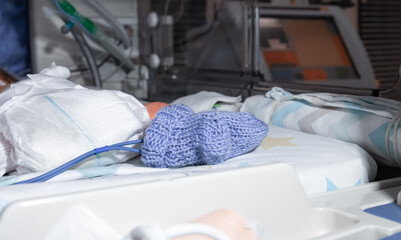 Premature newborn recovering in incubator. His foot in a blue sock in focus, with a premature infant pulse oximeter. Intensive care unit, ventilator for artificial lung ventilation as the background