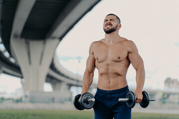 Fototapeta na wymiar Endurance and power concept. Motivated bearded European man raises barbells puts all efforts in lifting heavy weight clenches teeth poses with sport equipment under bridge, has muscular body
