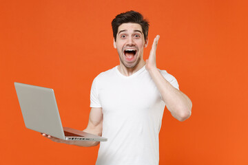 Surprised excited young man in basic blank white t-shirt standing working on laptop pc computer put...