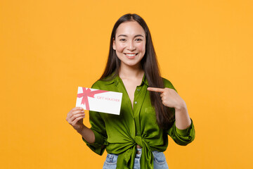 Smiling beautiful young brunette asian woman wearing basic green shirt standing pointing index finger on gift certificate looking camera isolated on bright yellow colour background, studio portrait.