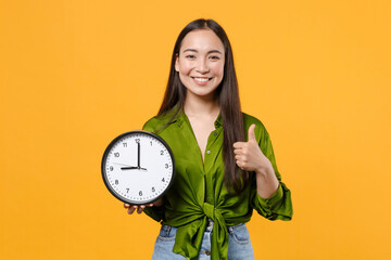 Smiling cheerful attractive young brunette asian woman 20s wearing basic green shirt standing hold clock showing thumb up looking camera isolated on bright yellow colour background, studio portrait.