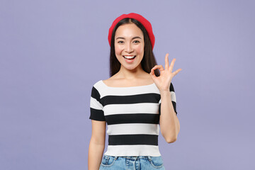 Obraz na płótnie Canvas Beautiful cheerful funny smiling young brunette asian woman wearing striped t-shirt red beret standing showing OK gesture looking camera isolated on pastel violet colour background studio portrait.