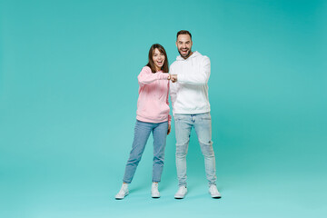 Full length of laughing funny young couple friends man woman in white pink casual hoodie holding hands folded giving fists bump looking camera isolated on blue turquoise background studio portrait.