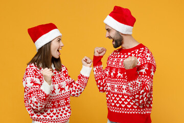 Happy young Santa couple friends man woman in sweater Christmas hat doing winner gesture looking at each other isolated on yellow background studio. Happy New Year celebration merry holiday concept.