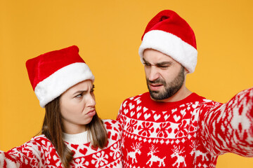 Close up offended young Santa couple friends man woman in red sweater, Christmas hat doing selfie shot on mobile phone isolated on yellow background. Happy New Year celebration merry holiday concept.