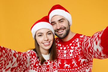 Close up smiling young Santa couple friends man woman in red sweater, Christmas hat doing selfie shot on mobile phone isolated on yellow background. Happy New Year celebration merry holiday concept.