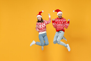 Full length of overjoyed young Santa couple friends man woman in red sweater Christmas hat jumping doing winner gesture isolated on yellow background. Happy New Year celebration merry holiday concept.