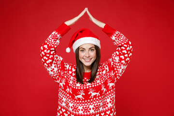 Smiling young brunette Santa woman in sweater, Christmas hat holding hands above head like roof of house isolated on red background, studio portrait. Happy New Year celebration merry holiday concept.