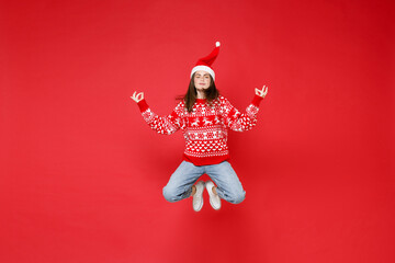 Full length of young brunette Santa woman in sweater, Christmas hat jumping hold hands in yoga gesture relaxing meditating isolated on red background. Happy New Year celebration merry holiday concept.