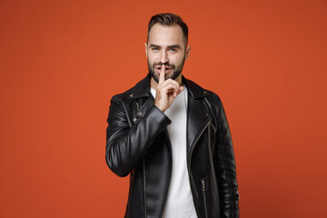 Secret young bearded man in basic white t-shirt black leather jacket standing saying hush be quiet with finger on lips shhh gesture looking camera isolated on orange colour background studio portrait.