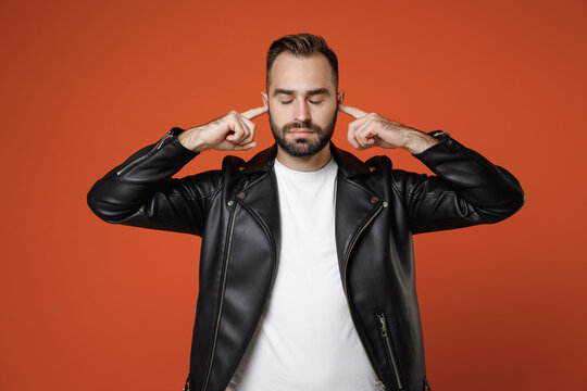 Calmed young bearded man 20s wearing basic white t-shirt black leather jacket standing covering ears with fingers keeping eyes closed isolated on bright orange colour wall background studio portrait.