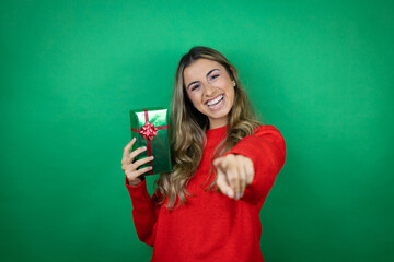 Young beautiful girl holding gift over isolated green background laughing at you, pointing finger to the camera with hand over body, shame expression