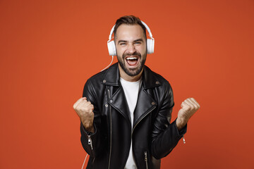 Joyful young bearded man in casual basic white t-shirt, black leather jacket standing listening music with headphones doing winner gesture isolated on bright orange colour background studio portrait.