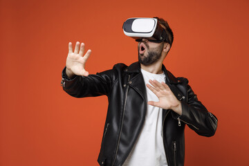 Shocked young bearded man 20s wearing white t-shirt black leather jacket standing watching in vr headset gadget touch something like push click on button isolated on orange background studio portrait.