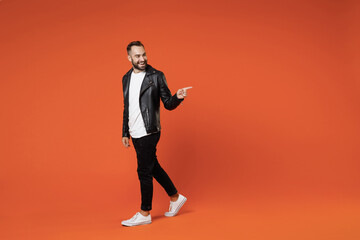 Fototapeta na wymiar Full length side view of smiling young bearded man in basic white t-shirt black leather jacket standing pointing index finger aside on mock up copy space isolated on orange background studio portrait.