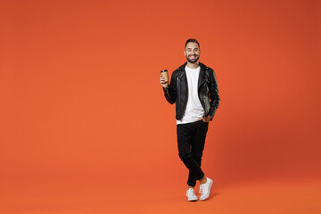 Fototapeta na wymiar Full length of smiling young bearded man wearing basic white t-shirt black leather jacket standing hold paper cup of coffee or tea looking camera isolated on orange colour background studio portrait.