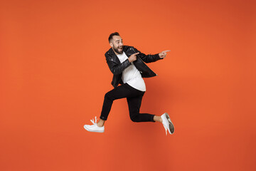 Fototapeta na wymiar Full length side view of excited surprised young bearded man 20s in basic white t-shirt black leather jacket jumping pointing index fingers aside isolated on orange colour background studio portrait.