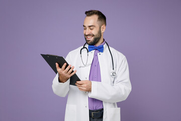 Smiling young bearded doctor man in white medical gown stethoscope hold clipboard with medical documents isolated on violet background studio portrait. Healthcare personnel health medicine concept.