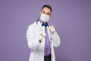 Young doctor man in medical gown stethoscope face mask safe from coronavirus virus covid-19 point index fingers on camera isolated on violet background. Healthcare personnel health medicine concept.