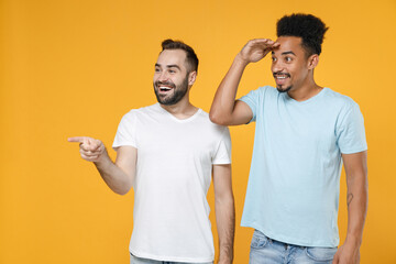 Funny young friends european african american men 20s in white blue t-shirts point index fingers aside holding hand at forehead looking far away distance isolated on yellow background studio portrait.