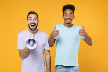 Excited cheerful young friends european african american men 20s in casual violet blue t-shirts screaming in megaphone showing thumbs up isolated on bright yellow colour background studio portrait.