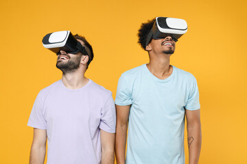 Funny cheerful funny young friends european african american men 20s wearing casual violet blue t-shirts watching in vr headset gadget isolated on bright yellow colour background studio portrait.