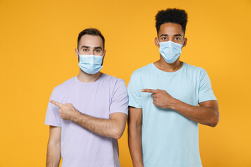 Young friends european african american men 20s in purple blue t-shirts sterile face mask to safe from coronavirus covid-19 pointing index fingers aside isolated on yellow background studio portrait.