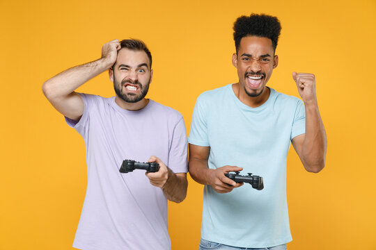 Displeased funny young friends european african american men 20s in violet blue t-shirts play pc game with joystick console doing winner gesture isolated on yellow colour background studio portrait.