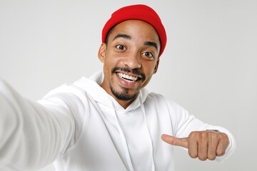 Close up of funny young african american man 20s wearing basic casual streetwear hoodie doing selfie shot on mobile phone pointing thumb on himself isolated on white wall background, studio portrait.