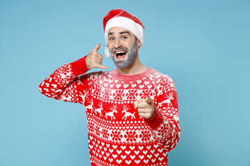 Excited Northern bearded man frozen face in Santa hat Christmas sweater doing phone gesture says call me back point on camera isolated on blue background. Happy New Year holiday winter time concept.
