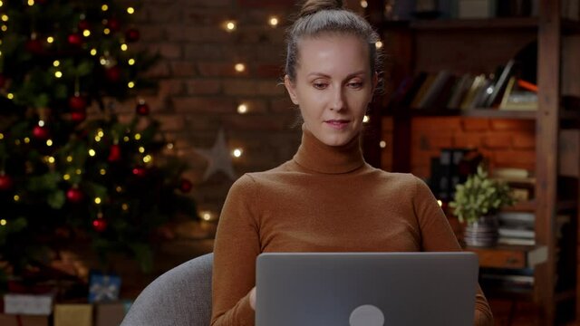White woman sitting at home in front of Christmas tree shopping online paying on laptop with bank card.