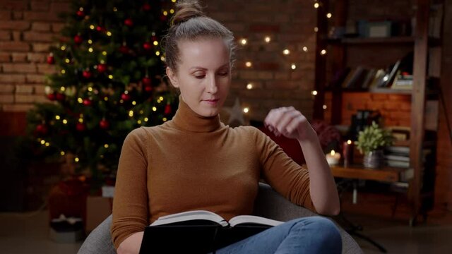 Adult woman reading book at home at Christmas in winter, dark room with Christmas tree.
