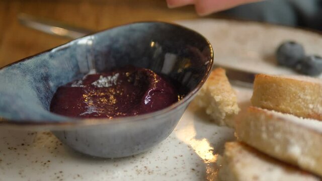 Cheesecake piece dipping in blue sauce bowl with delicious bright purple berry jam in vegan cafe extreme closeup slow motion