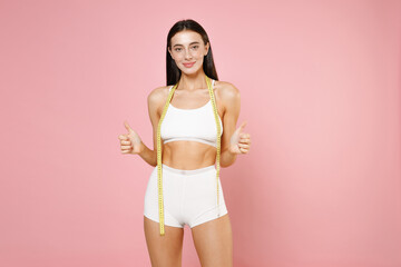 Fototapeta na wymiar Smiling young brunette woman 20s in white underwear with strong fit body posing hold measure tape showing thumbs up looking camera isolated on pastel pink colour wall background, studio portrait.