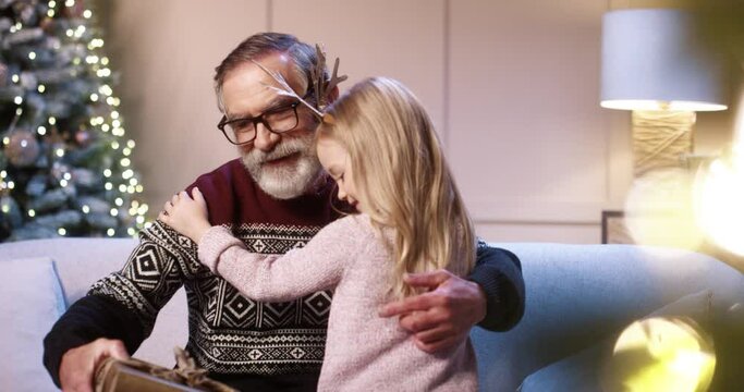 Close up portrait of happy Caucasian cute little girl giving present to old grandpa while sitting at cozy decorated home near glowing new year tree. Grandfather receiving xmas gift from grandchild