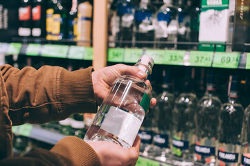 A male shopper in a supermarket holds a bottle of vodka in his hands. - obrazy, fototapety, plakaty