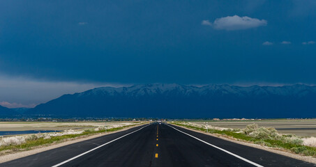 Straight road going towards a very stormy and black sky with the mountains on the horizon under heavy rain.  Angtelope island, Utah, United States of America