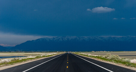 Straight road going towards a very stormy and black sky with the mountains on the horizon under...