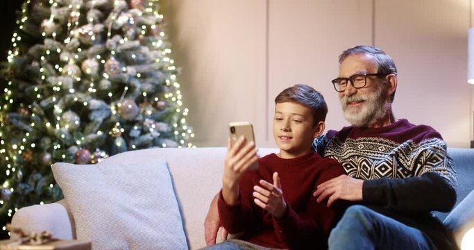 Portrait of happy smiling Caucasian old grandpa with teen grandchild sitting in decorated room and taking selfie photos on smartphone at home near glowing new year tree. Christmas holidays concept