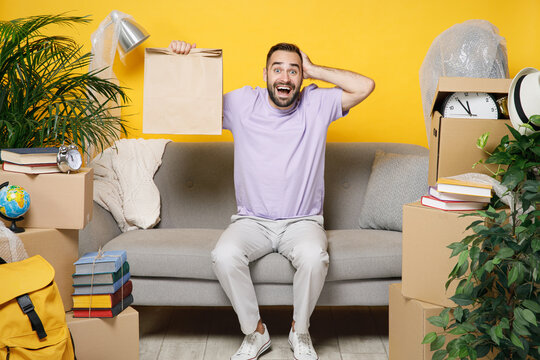 Excited owner man hold craft paper bag for takeaway sits in living room on sofa at home household unpacking stuff indoor rents flat isolated on yellow wall. Relocation moving in new apartment concept.