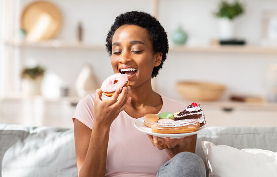 Young black woman on diet having cheat meal day, holding plate of sweets, stuffing her mouth with donut at home