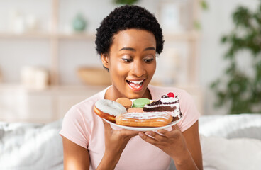 Diet breakdown and unhealthy nutrition concept. Young African American lady holding plate of sweet pastries at home