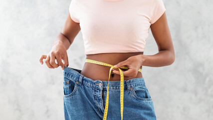 Black woman in old big jeans measuring her waist, showing results of slimming diet or liposuction, grey background - Powered by Adobe