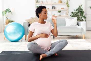 Keep hydrated during pregnancy. Black expectant lady having glass of water while exercising on yoga mat at home