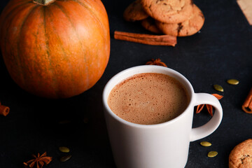 Cup of coffee with milk foam, spices, orange pumpkin, seeds, cookies on black table. Autumn drink concept. Fall, spicy latte, home, pastry, thanksgiving, coffee shop menu, closeup